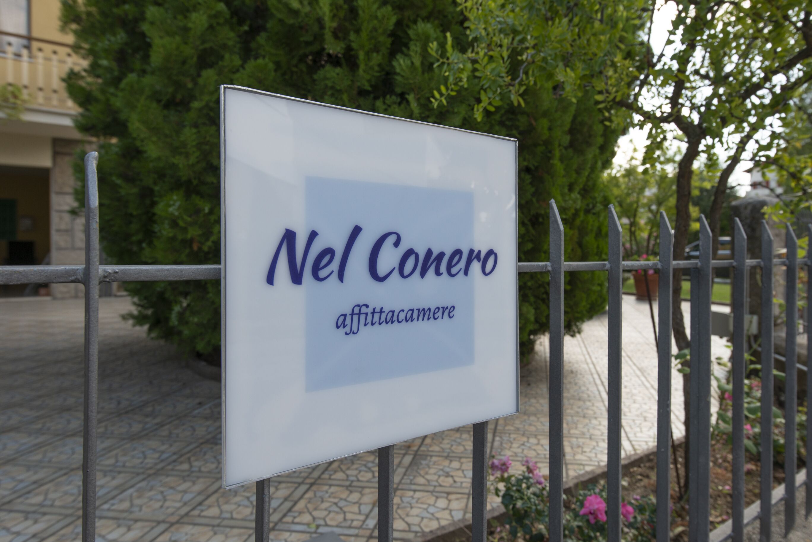 Ein Schild mit dem Schriftzug "Nel Conero affittacamere" vor einem Gebäude mit einem gepflasterten Eingang und einem Metallzaun.