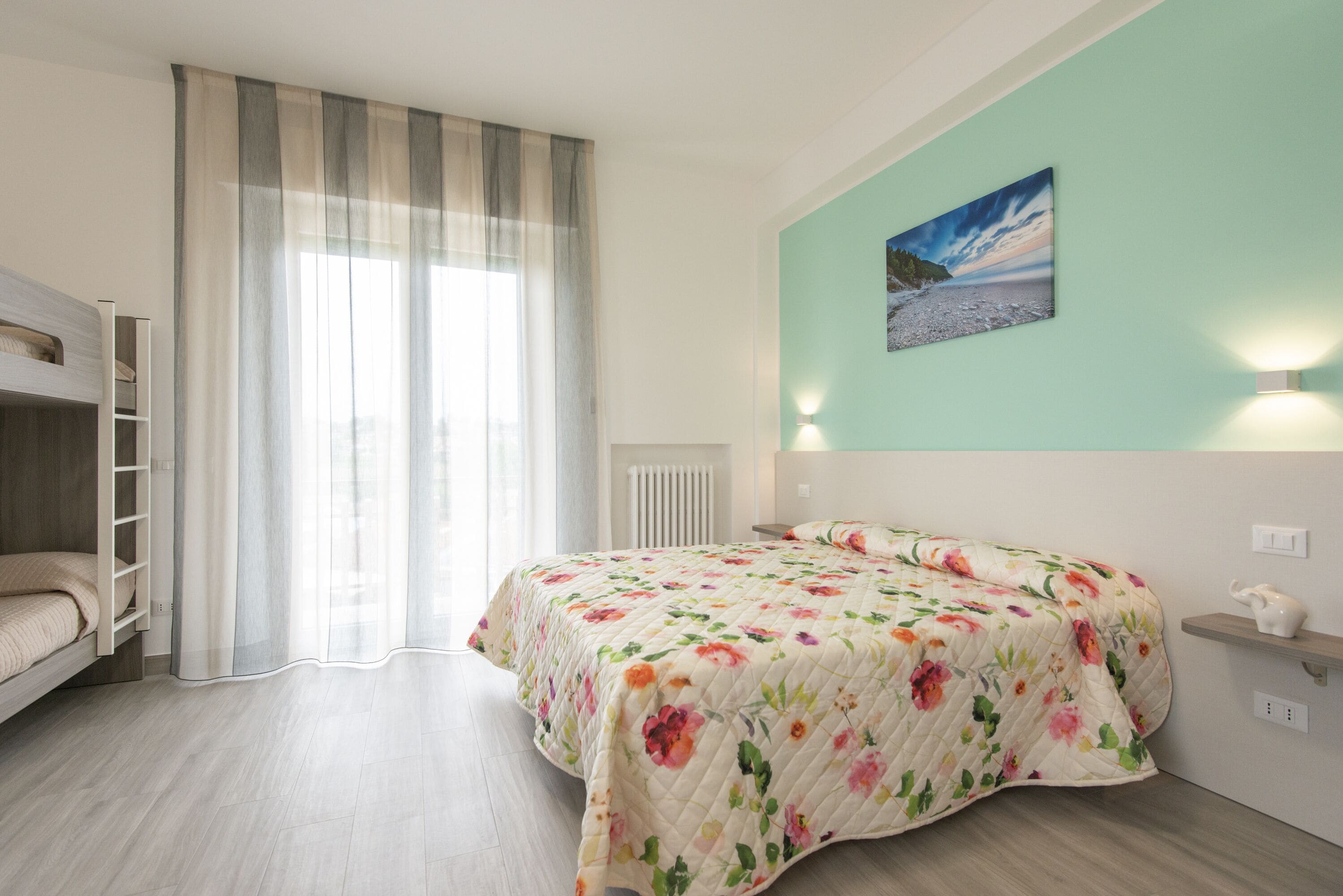 Un dormitorio con cama, ventana con cortinas, radiador, estante en la pared y un cuadro.