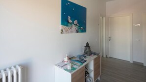 Une pièce avec un radiateur blanc, un meuble en bois contenant des magazines et un tableau représentant un paysage côtier.