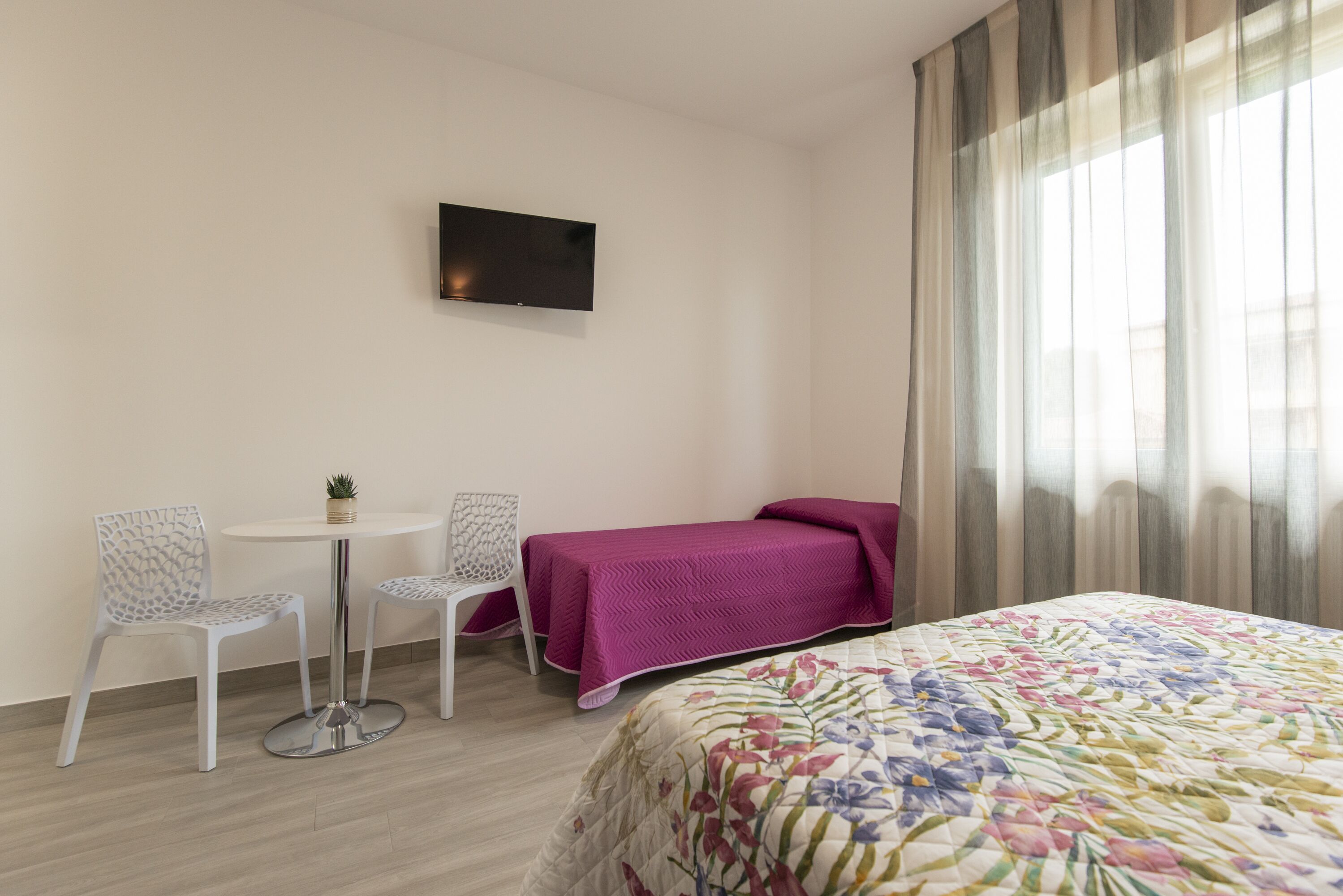 A hotel room with a bed, a small table with two chairs, a TV on the wall, and a window with curtains.