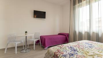 A hotel room with a bed, a small table with two chairs, a TV on the wall, and a window with curtains.