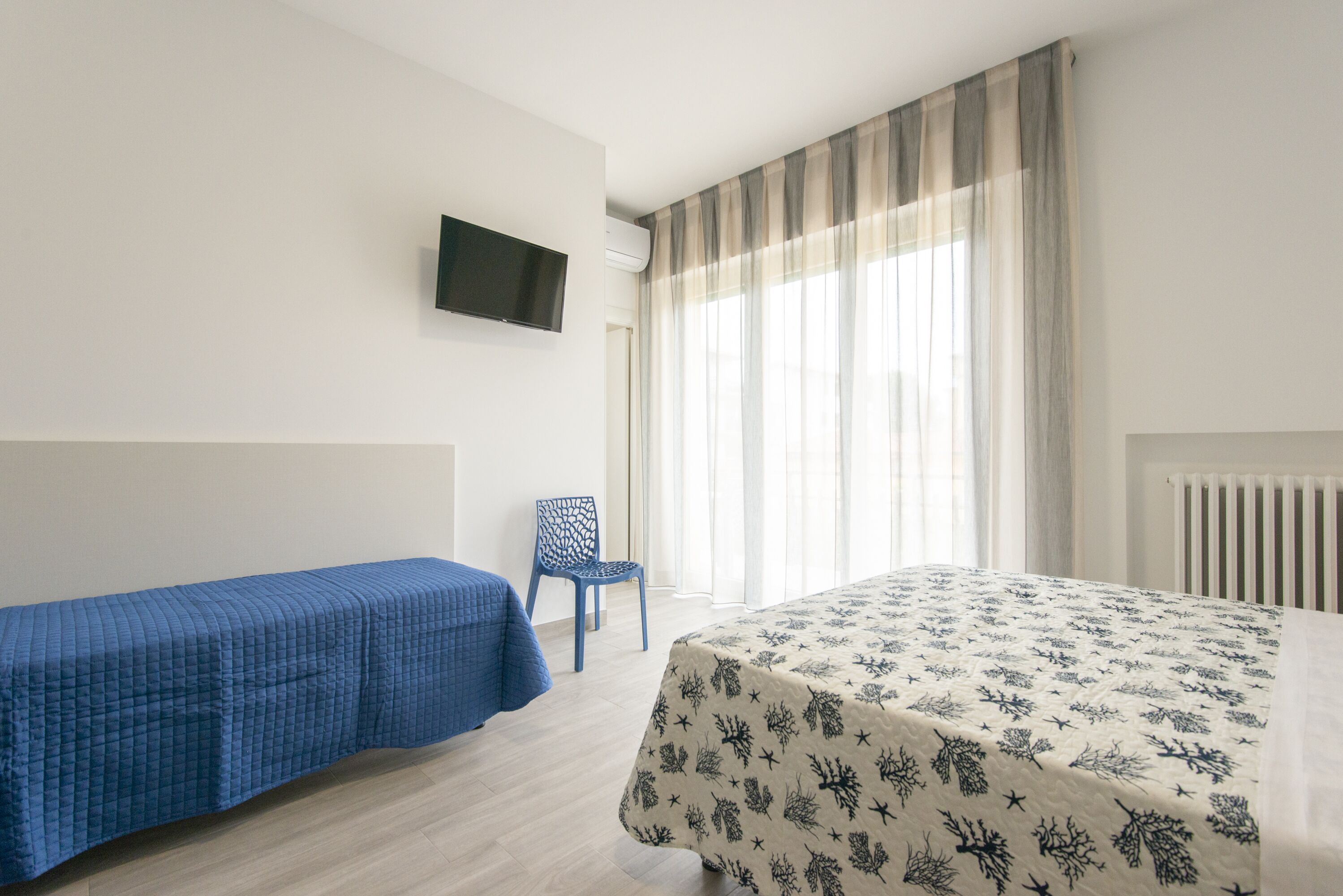 A hotel room with a bed, a chair, a TV mounted on the wall, and a window with curtains.