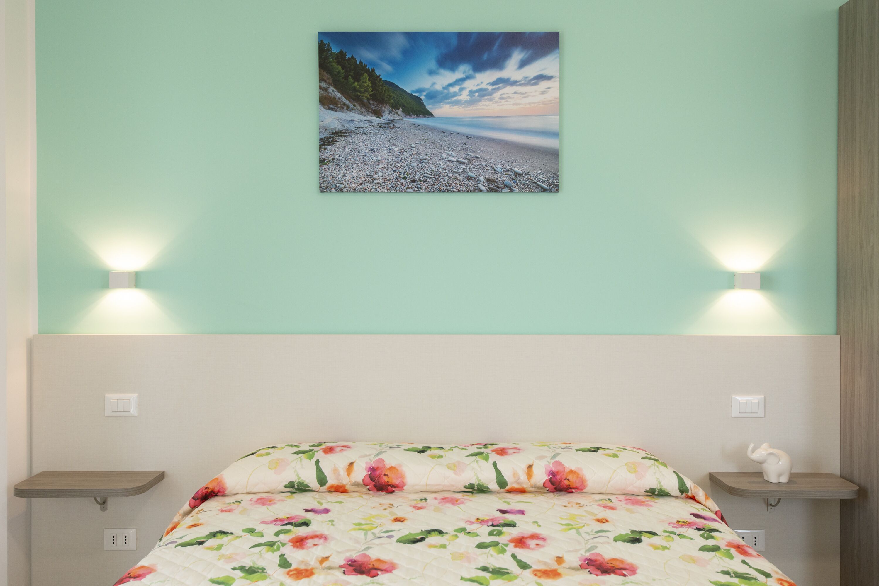 Un dormitorio con cama, ropa de cama floral, un cuadro colgado en la pared de una playa y lámparas de noche.
