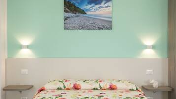 Un dormitorio con cama, ropa de cama floral, un cuadro colgado en la pared de una playa y lámparas de noche.