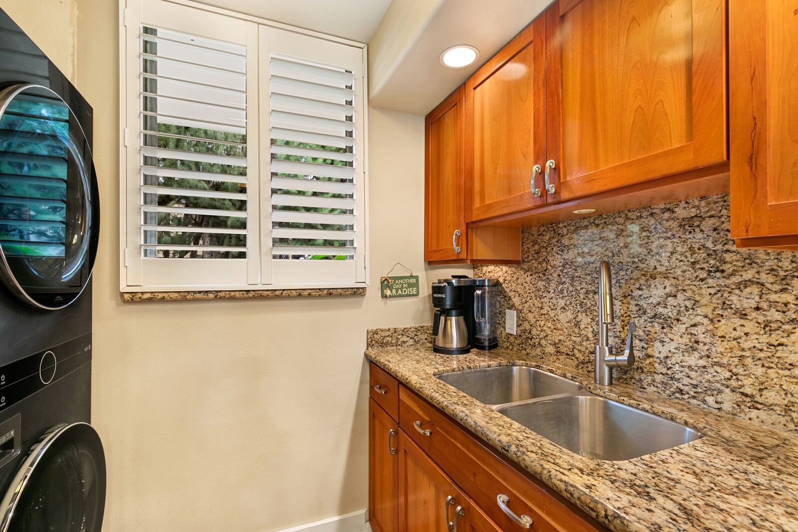 A kitchen with wooden cabinets, a granite countertop, a sink, a coffee maker, and a window with blinds.