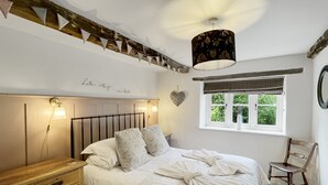 Un dormitorio con un aparador de madera, una cama con ropa de cama blanca, una lámpara colgante, una ventana con vista a follaje y una decoración con forma de corazón en la pared.