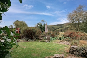 Un jardin avec une table et des chaises, entouré de verdure et avec des collines en arrière-plan.