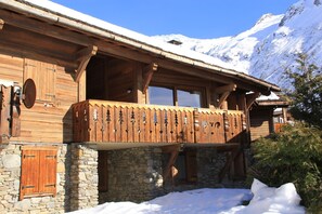 Un chalet en bois doté d’un balcon, de murs en pierre et surplombant un paysage montagneux enneigé.