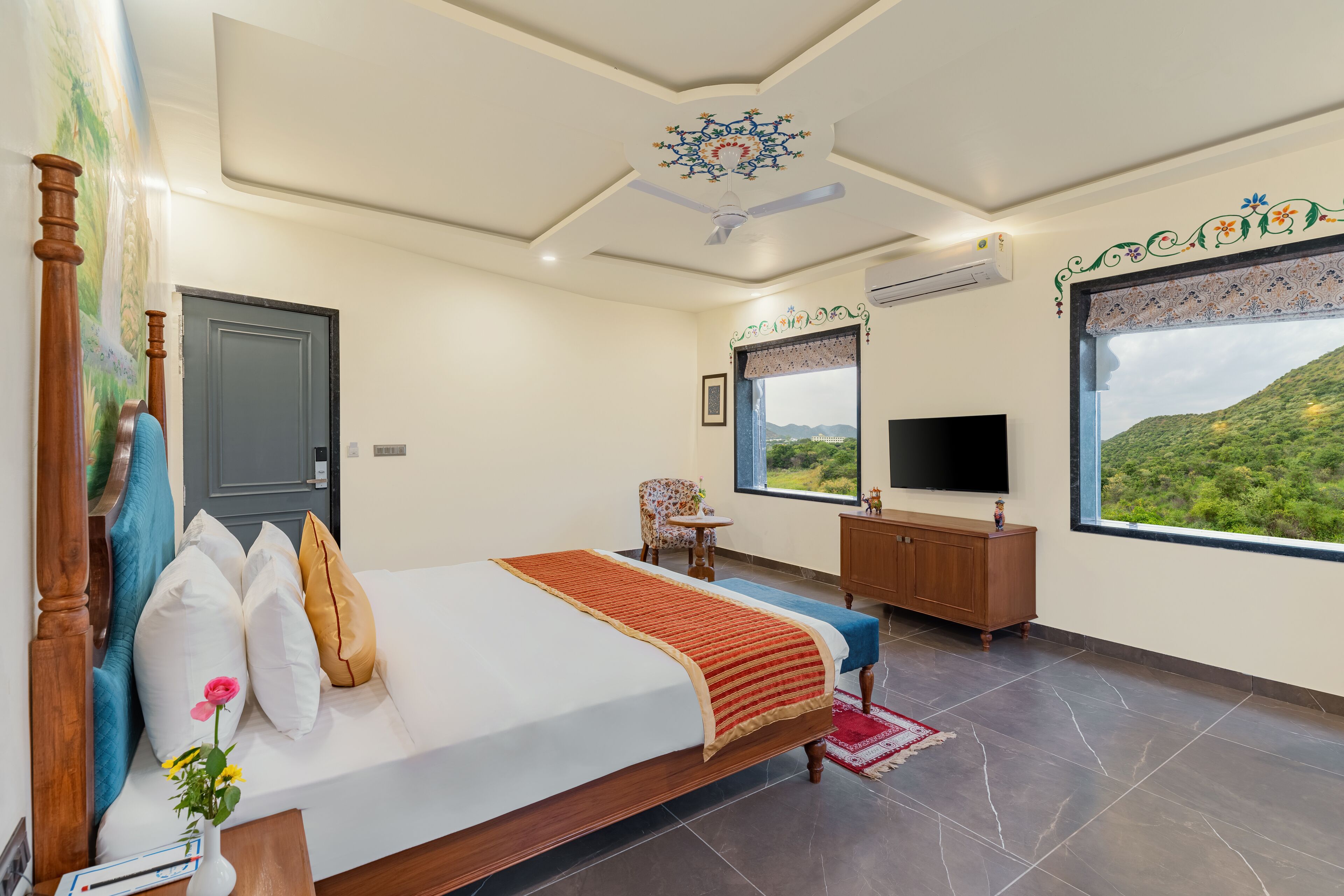 Quarto de hotel com cama, televisão, janela vista para vegetação e ventilador de teto.