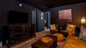 A dark-themed living room with a large flat-screen TV, a wooden coffee table, a sofa with patterned cushions, and a decorative lamp. - walk to square! Cozy 2 BR bungalow, moody design (Dahlonega)