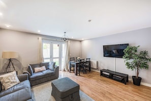 A modern living room with a grey sofa, ottoman, dining table, and chairs. A flat-screen TV is mounted on the wall, and a potted plant is on the floor.