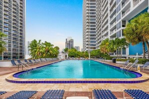 Une piscine entourée de chaises longues et de palmiers, avec de grands immeubles en arrière-plan.