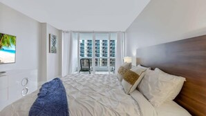 A hotel room with a bed, a TV, a window with a view of buildings, and a chair by the window.