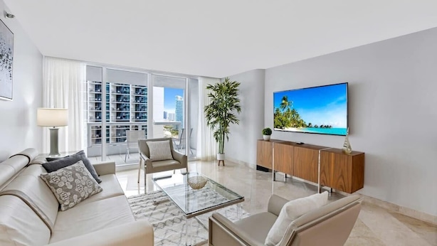 A modern living room with a sofa, armchairs, a coffee table, a TV, and a large window with a city view.