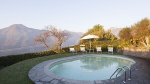 A swimming pool with a stone border, surrounded by grass and a hedge. There are lounge chairs and an umbrella. Mountains are visible in the background. - Casa Pedona (Camaiore)