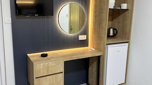 A compact room with a dark blue wall, a wooden desk with drawers, a small white refrigerator, a mounted TV, and a round mirror. - Enda Lara Hotel (Antalya)