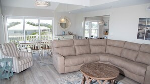A spacious living area with a large beige sectional sofa, a round wooden coffee table, and a striped armchair. A dining table with chairs is visible in the background, and a large window offers a view of the outdoors. - Come feel the "SEA BREEZE" - Minimum 2 week rental starting on Sundays (Emerald Isle)