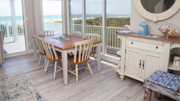 A dining area with a wooden table and chairs, a white cabinet, and a view of the ocean through large windows. - Come feel the "SEA BREEZE" - Minimum 2 week rental starting on Sundays (Emerald Isle)