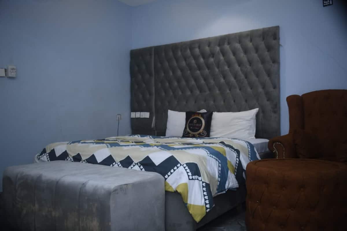 Une chambre à coucher avec une tête de lit capitonnée, un lit avec une literie à motifs, un fauteuil marron et un pouf gris.