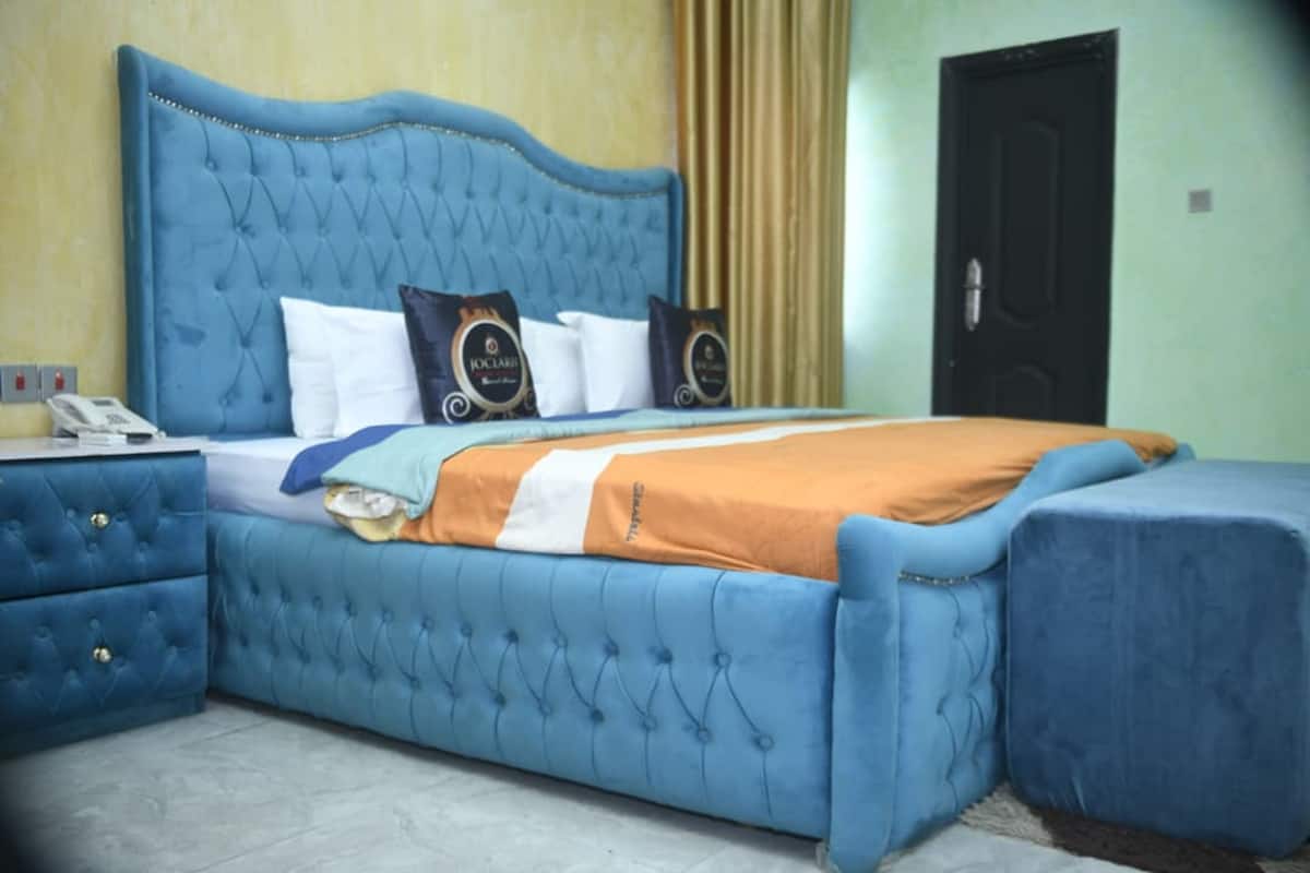 Une chambre à coucher avec un lit recouvert d’un tissu bleu, une table de chevet avec un téléphone, et une porte visible au fond.