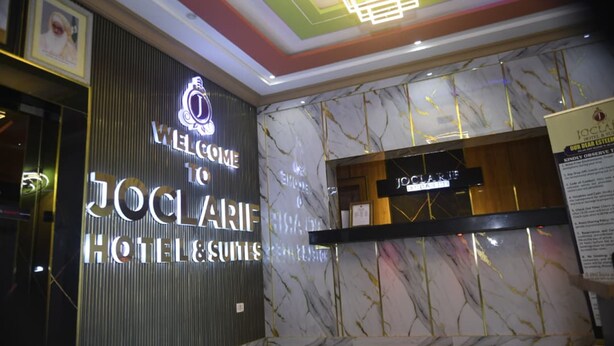 Un hall d’accueil d’hôtel avec un mur en marbre, un comptoir de réception et une enseigne sur laquelle on peut lire : « BIENVENUE À L’HÔTEL JOCLAIRE & SUIRES ».