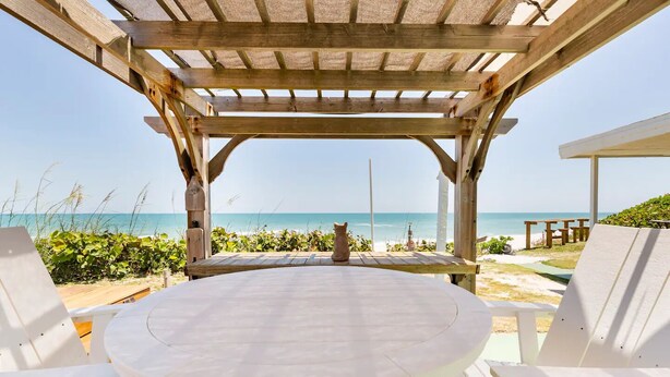 A wooden pergola with a round table and white chairs overlooking a beach. - Charming Two Story House located directly on Melbourne Beach. (Melbourne Beach)