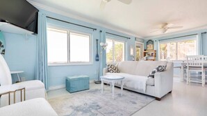 A spacious living room with a white sofa, a blue ottoman, and a small round table. There are large windows allowing natural light, and a ceiling fan is visible. - Charming Two Story House located directly on Melbourne Beach. (Melbourne Beach)