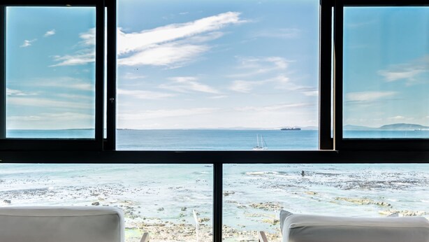 A room with a large window overlooking a rocky beach and the sea. There are two chairs and a small table with a bowl. - Magical Apartment w/Uninterrupted Ocean Views (Mouille Views) (Cape Town)