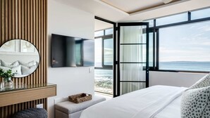 A bedroom with a large bed, a TV mounted on the wall, a window with a view of the sea, and a wooden panel wall. - Magical Apartment w/Uninterrupted Ocean Views (Mouille Views) (Cape Town)