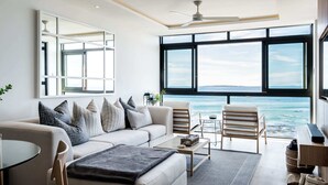 A modern living room with a large window overlooking the sea, a ceiling fan, and a minimalist design. - Magical Apartment w/Uninterrupted Ocean Views (Mouille Views) (Cape Town)