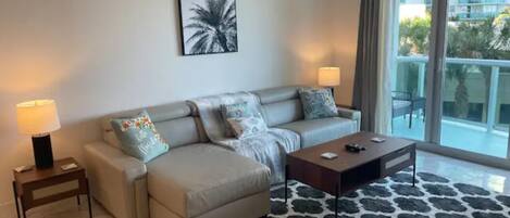 Relax in this spacious living room with a large sofa, side table, and coffee table, offering a view of the surrounding buildings and greenery.
