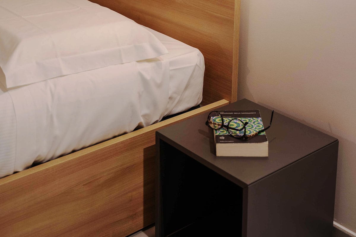 A neatly made bed with a wooden headboard, a bedside table with a book and glasses, and a wall-mounted light fixture.