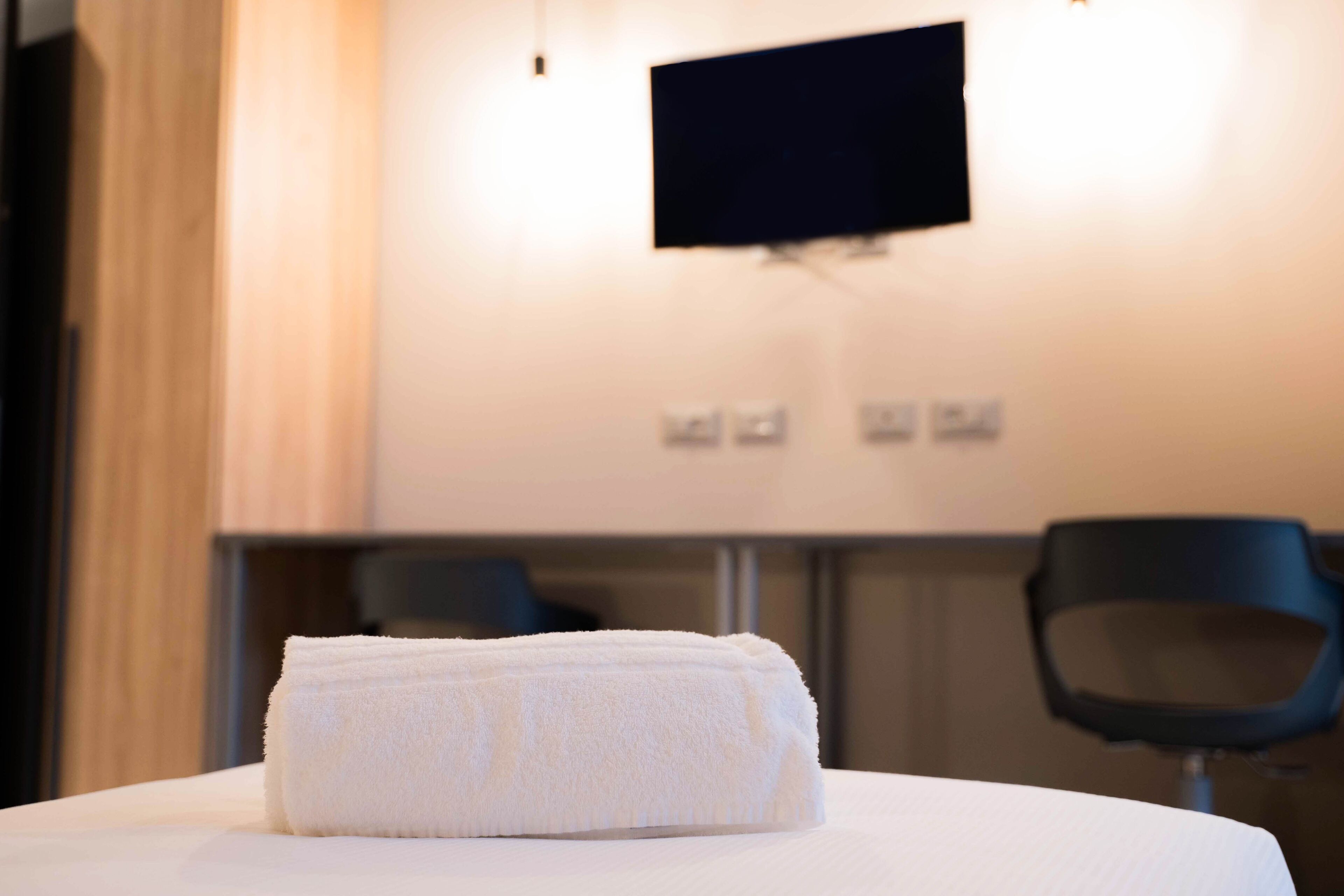 Una cama bien hecha con una toalla blanca doblada, un televisor de pantalla plana montado en la pared y una silla al fondo.