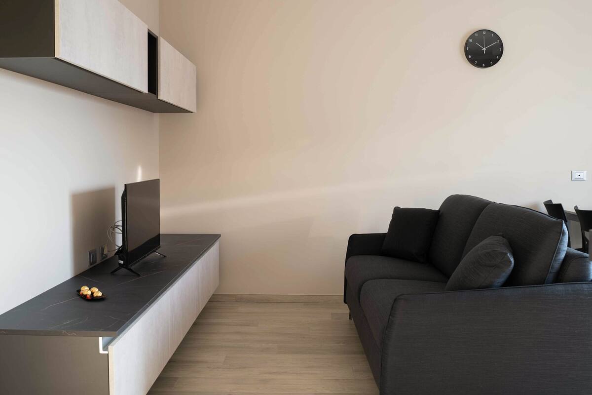 A modern living room with a flat-screen TV on a dark counter, a dark sofa with cushions, and a wall clock.