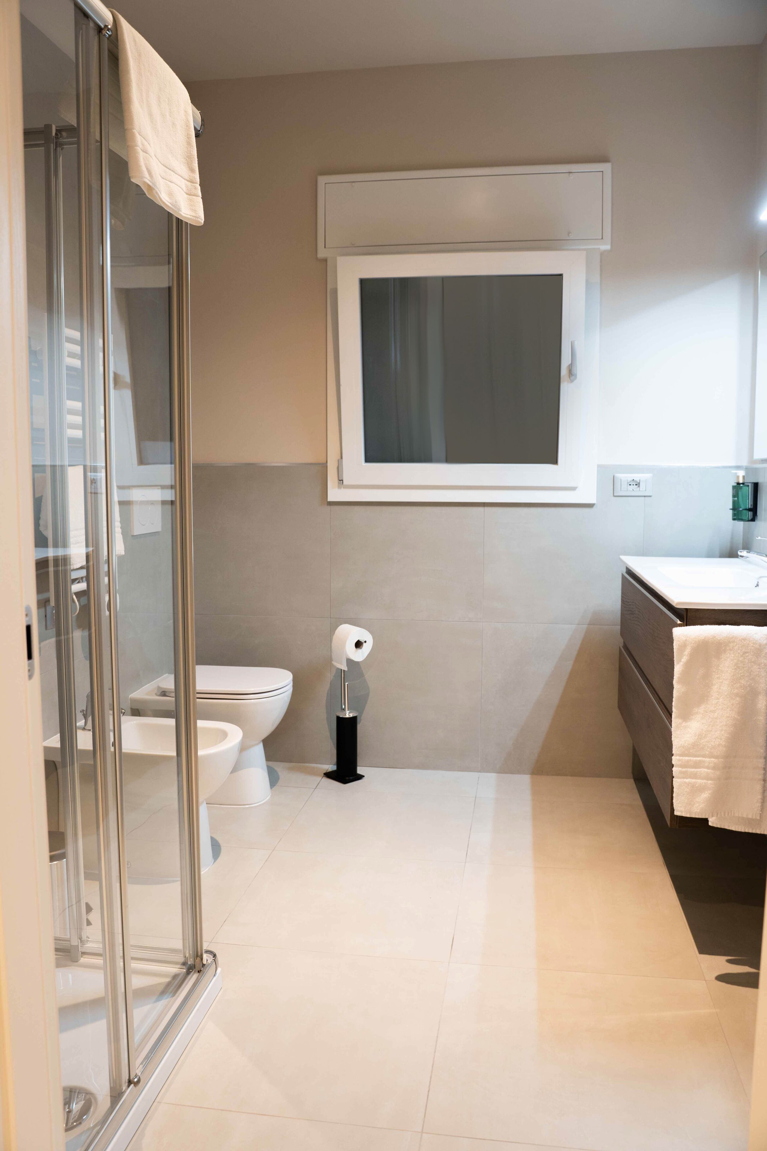 A modern bathroom with a glass shower enclosure, a toilet, a sink, and a window above the mirror.