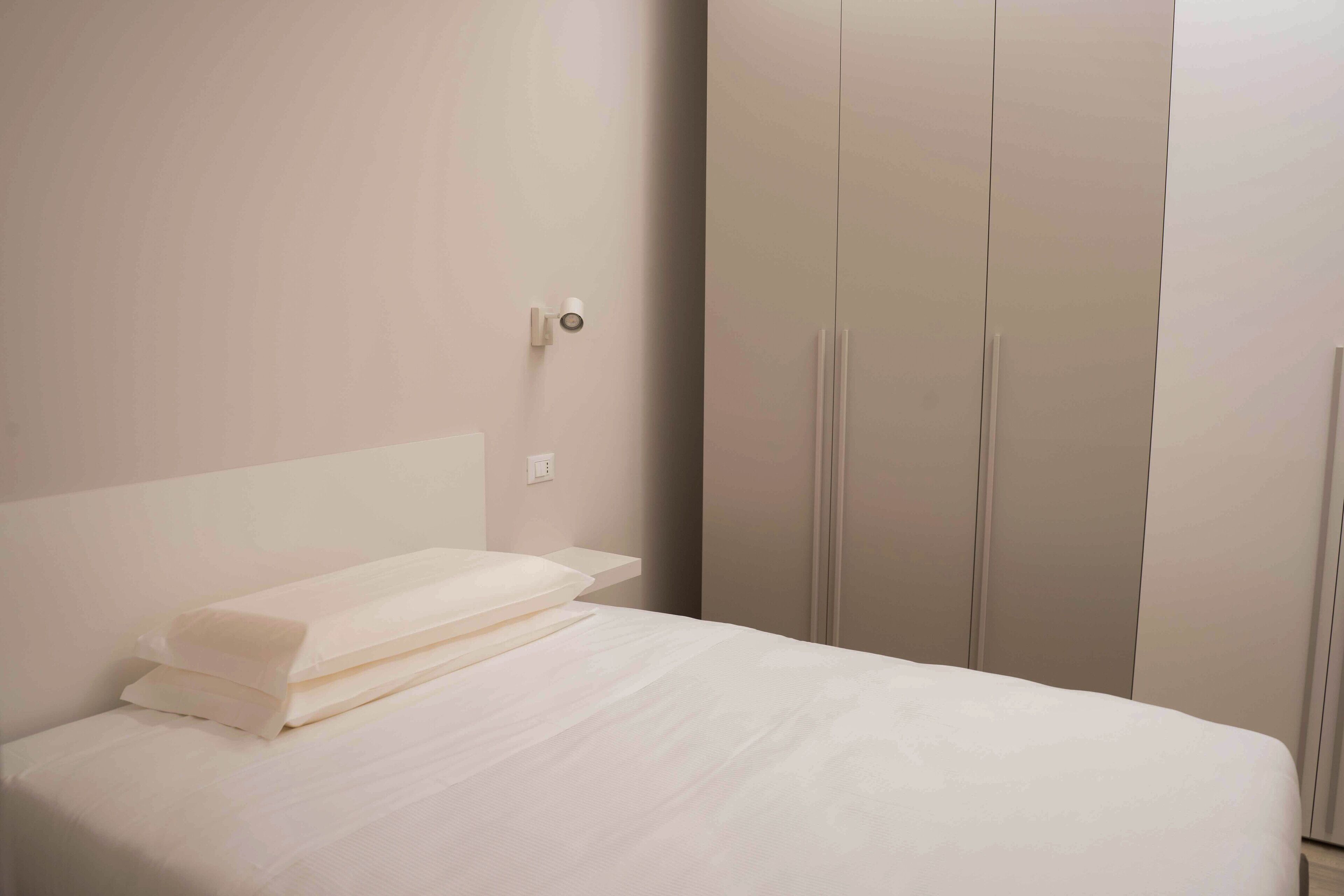 A neatly made bed with white linens in a room with a minimalist design, featuring a large wardrobe and a wall-mounted light fixture.