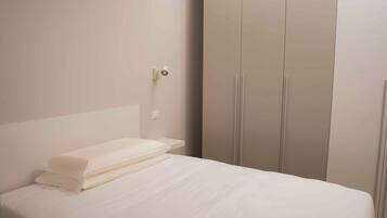 Ein ordentlich bezogenes Bett mit weißen Leinen in einem minimalistisch eingerichteten Zimmer, mit großem Kleiderschrank und Wandleuchte.
