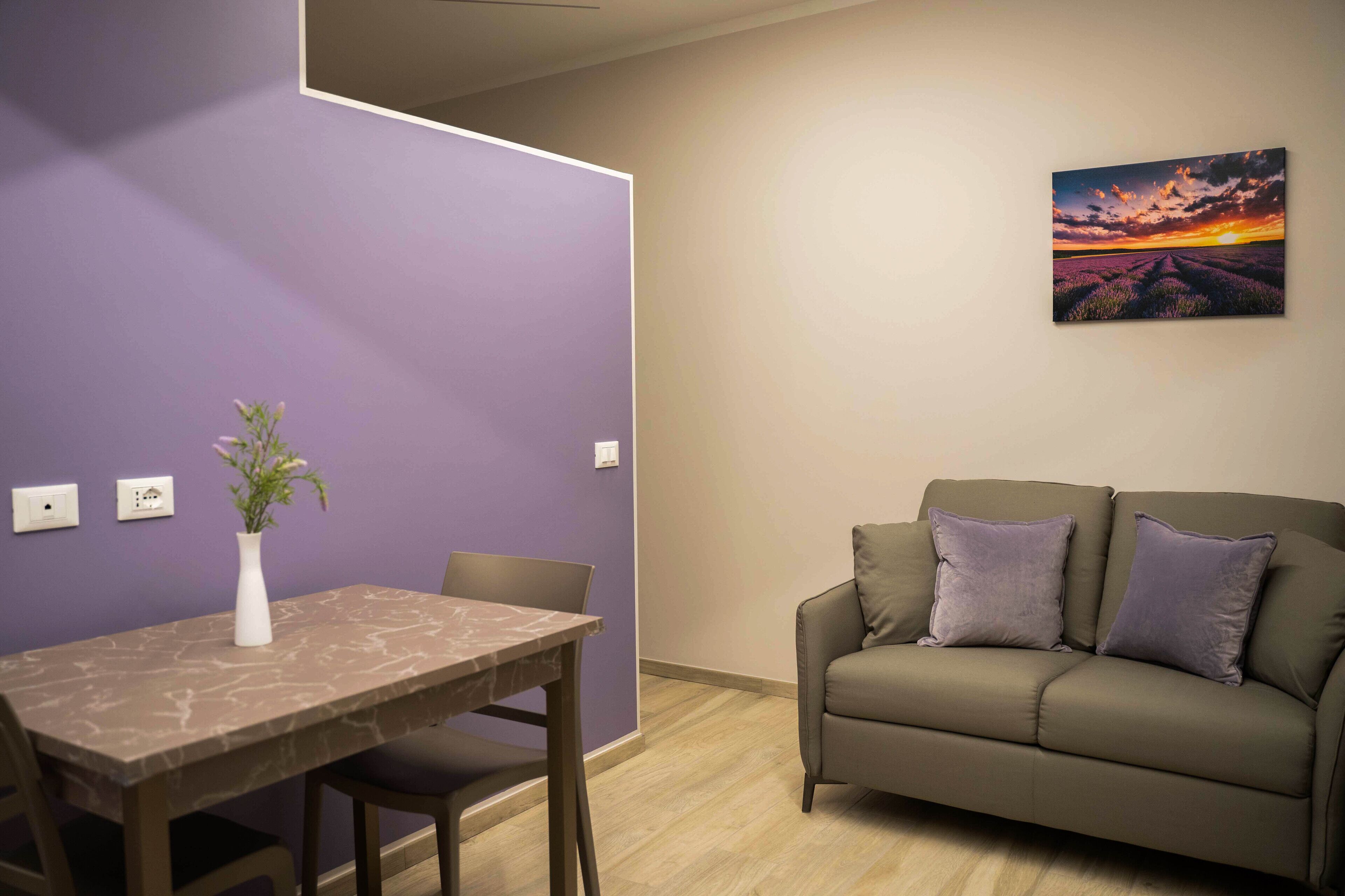 A room with a dining table, chairs, a vase with flowers, a sofa with cushions, and a painting on the wall.