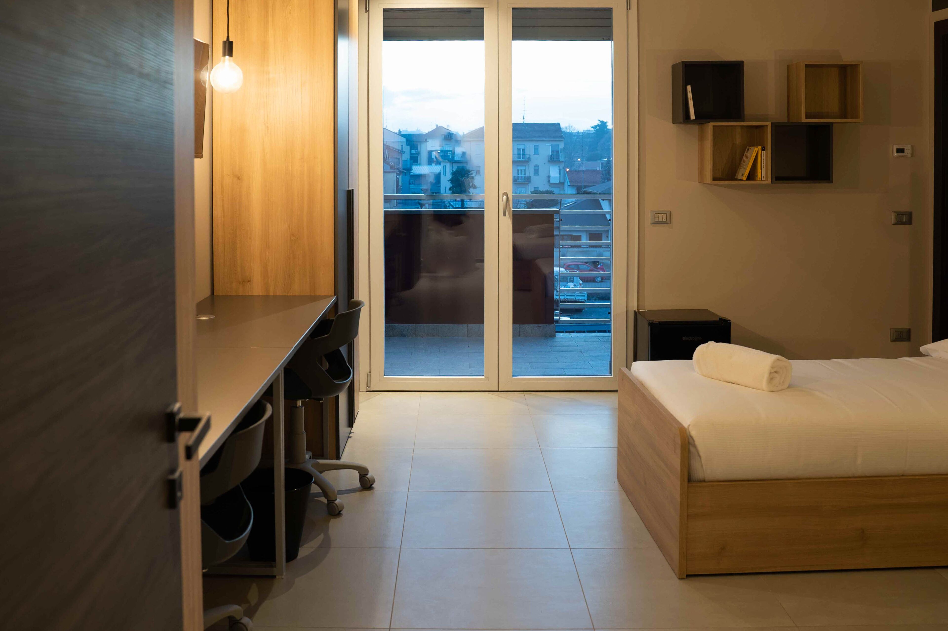 Una habitación de hotel moderna con una cama de madera, un escritorio con una silla, un televisor y un ventanal corredero que da a un balcón con vistas a la ciudad.