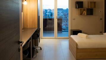 Una habitación de hotel moderna con una cama de madera, un escritorio con una silla, un televisor y un ventanal corredero que da a un balcón con vistas a la ciudad.