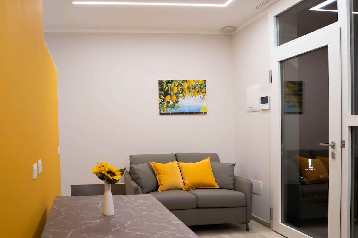 A modern living room with a grey sofa, yellow cushions, a vase with sunflowers, and a painting of a landscape.