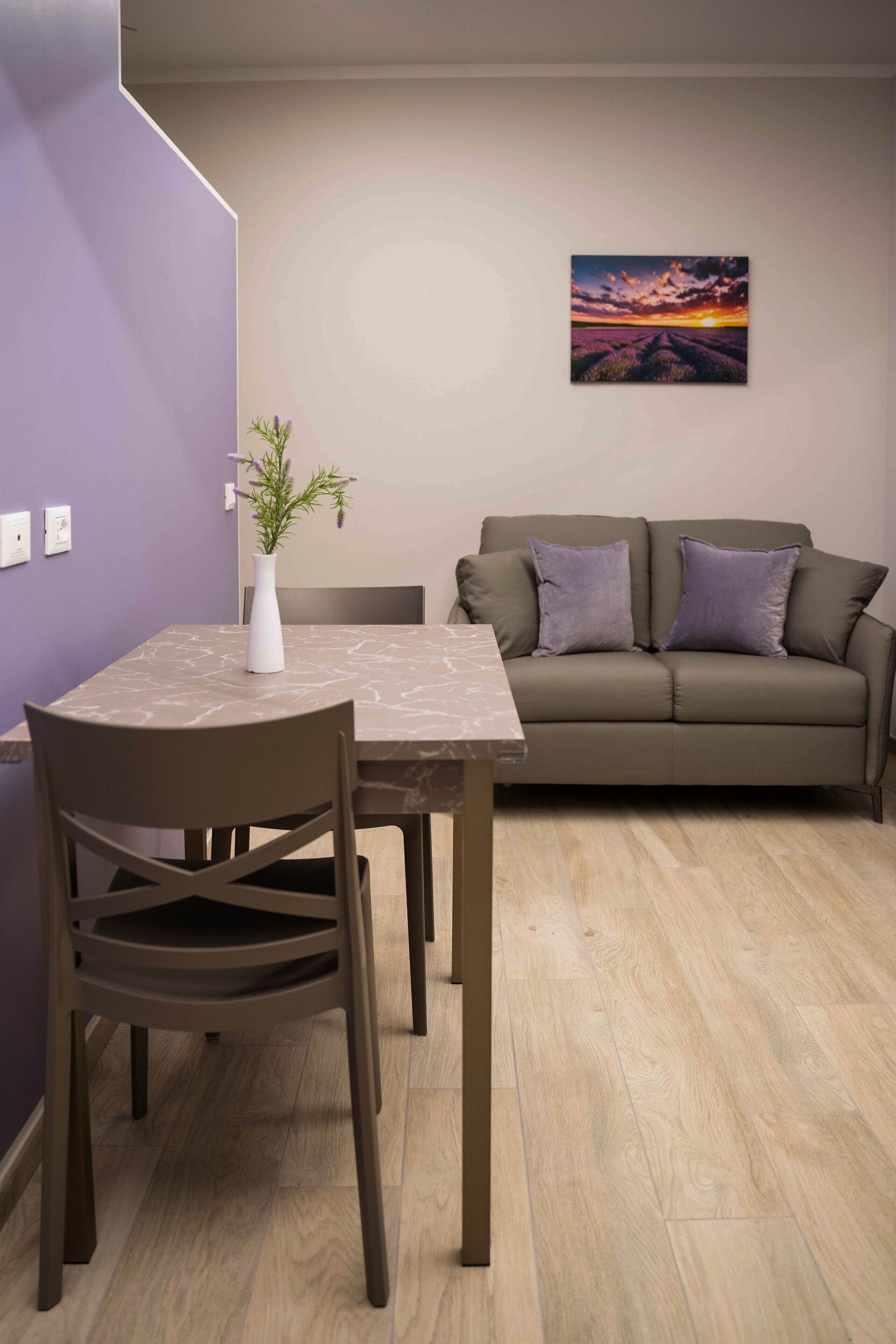 Un coin repas avec une table et des chaises, un canapé avec des coussins, un vase avec une plante et un tableau représentant un coucher de soleil accroché au mur.