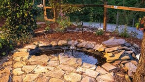 A stone fountain surrounded by a wooden fence, with a garden area and a wooden deck in the background. - 40 Acres w/Waterfalls, Vineyard, Stream, 5 bedroom (Moravian Falls)