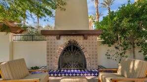 A patio with two striped lounge chairs, a patterned tiled fireplace, and a view of palm trees. - Perfect View, Convenient and Peaceful King Casita at Legacy Villas (La Quinta)