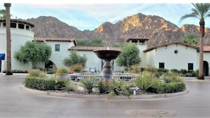 A courtyard with a central fountain, surrounded by buildings with Mediterranean-style architecture, set against a mountainous backdrop.