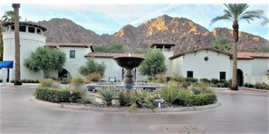A courtyard with a central fountain, surrounded by buildings with Mediterranean-style architecture, set against a mountainous backdrop.