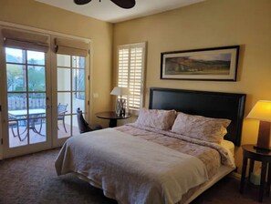 Relax in this serene space with a large bed, a window with blinds, and a sliding glass door leading to an outdoor area with a table and chairs.