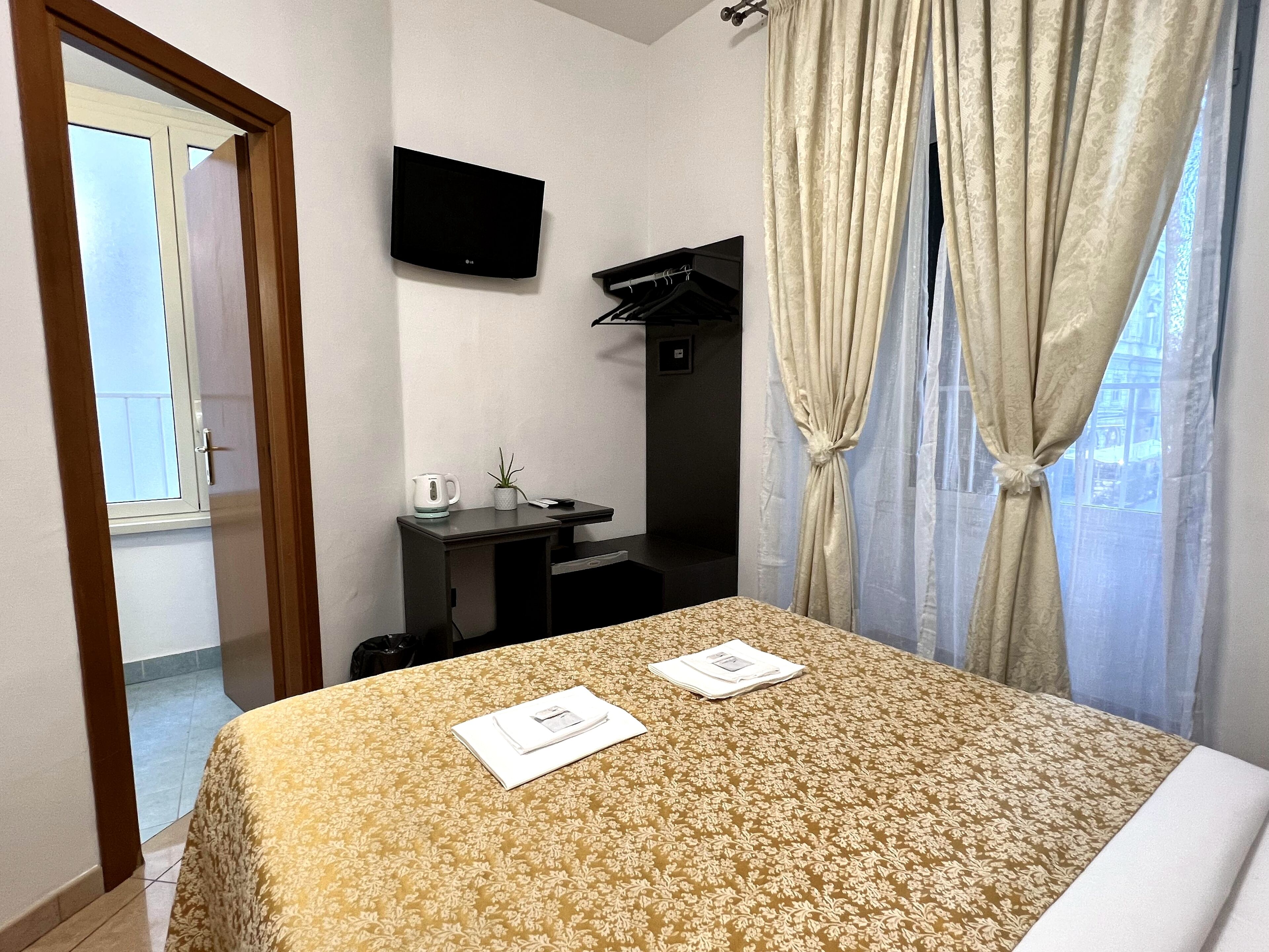 Una habitación de hotel con una cama, un televisor montado en la pared, una mesita con un hervidor y una planta, y un armario.