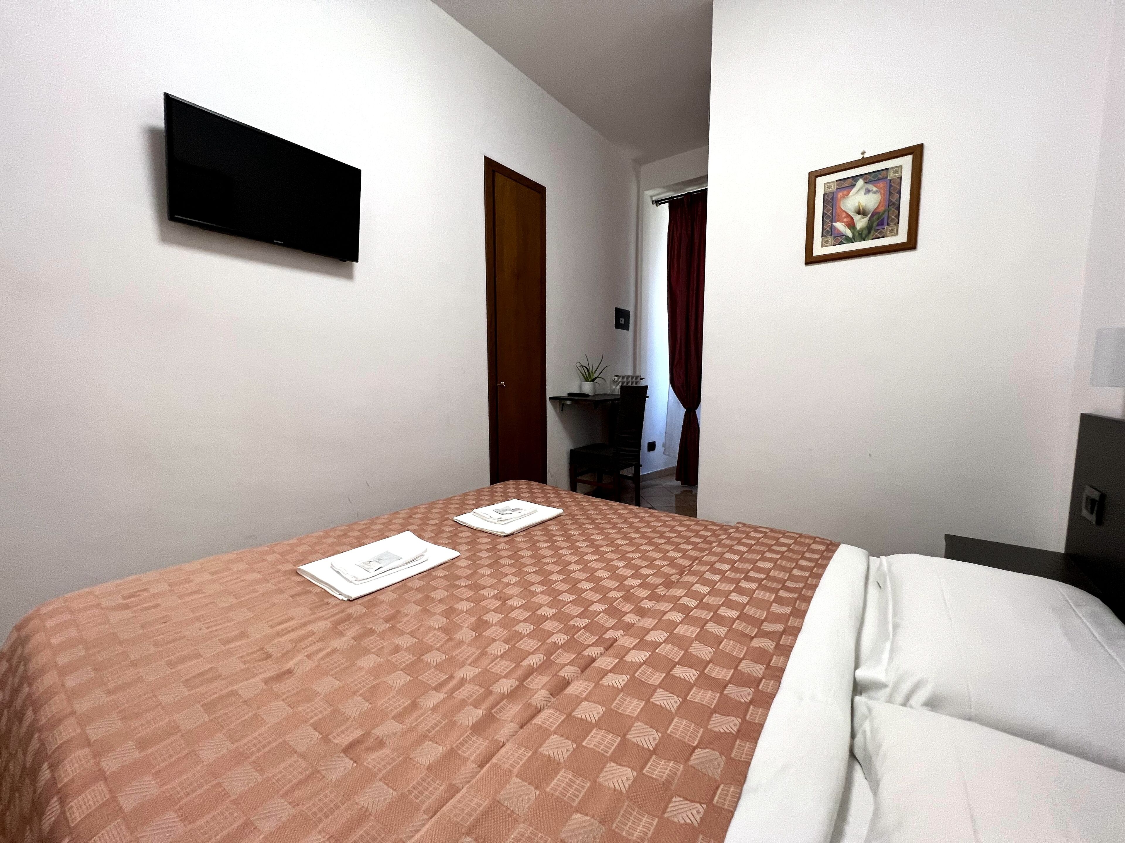 A hotel room with a bed, a flat-screen TV mounted on the wall, a small desk with a chair, and a framed picture on the wall.