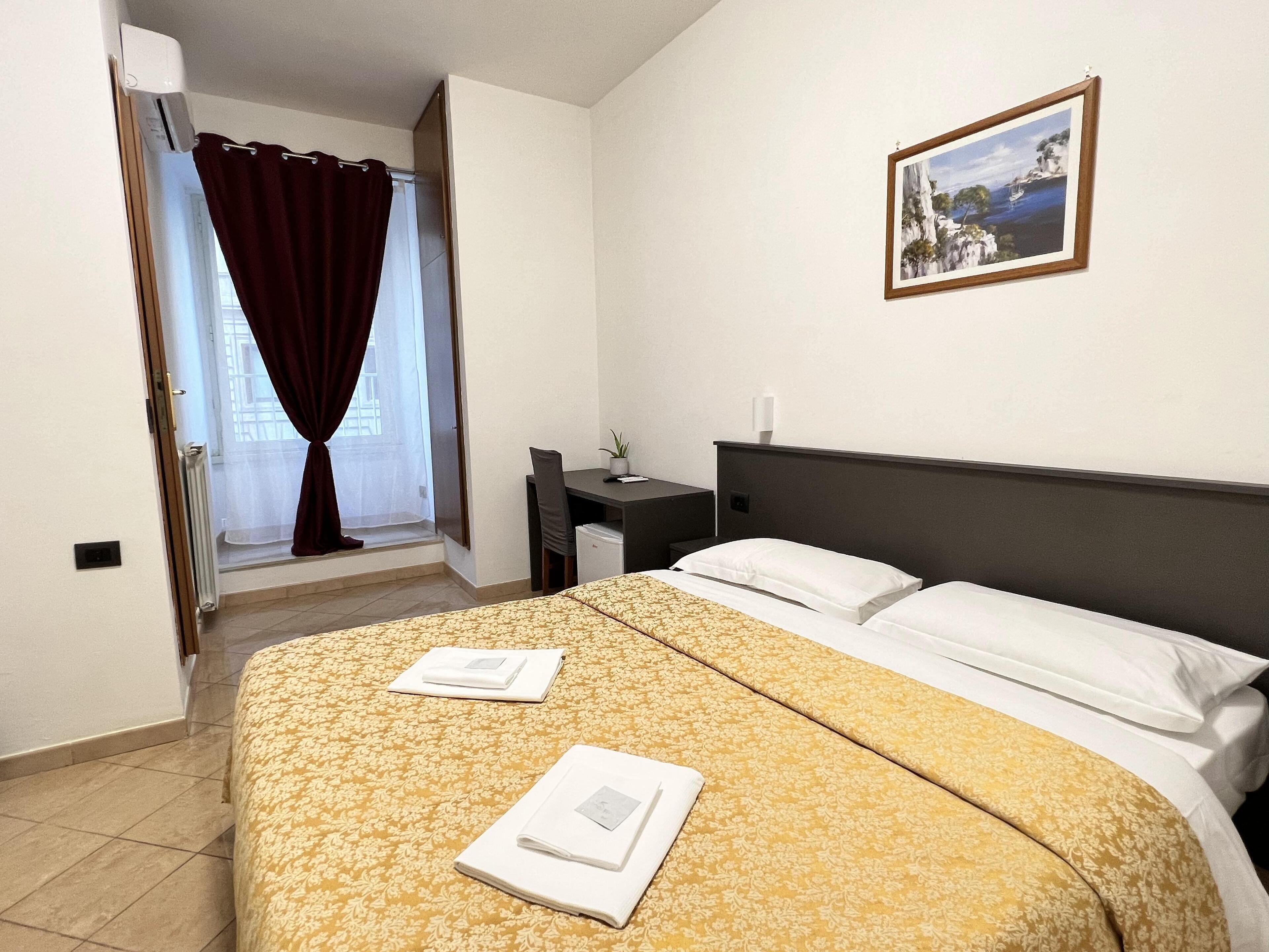 A hotel room with a bed, a desk, a chair, a window with curtains, and a framed picture on the wall.